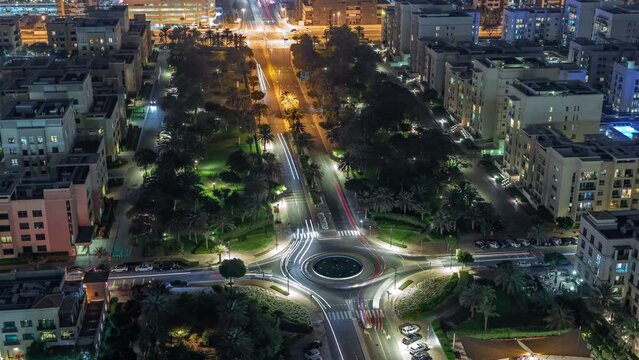 Traffic on a road with roundabout and low rise buildings in Greens district aerial night timelapse. Dubai skyline with palms and trees