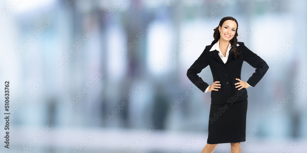 Wall mural Portrait of happy smiling businesswoman with hands on hips, copy space area for some text. Confident woman in black suit standing on blurred office background. Business concept.