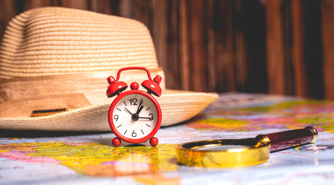 Tempo de f&eacute;rias. Um rel&oacute;gio despertador vermelho sobre um mapa do mundo com uma lupa e um chap&eacute;u de sol feminino na composi&ccedil;&atilde;o da imagem. Conceito de planejamento de f&eacute;rias e viagem ao exterior.