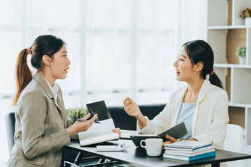 Young beautiful asian business woman is talking to her colleague, while sitting at the desk in a modern office. Concept of business success.