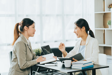 Obraz premium Young beautiful asian business woman is talking to her colleague, while sitting at the desk in a modern office. Concept of business success.