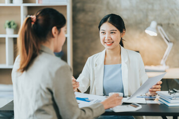Young beautiful asian business woman is talking to her colleague, while sitting at the desk in a modern office. Concept of business success.