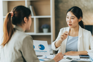 Obraz premium Young beautiful asian business woman is talking to her colleague, while sitting at the desk in a modern office. Concept of business success.