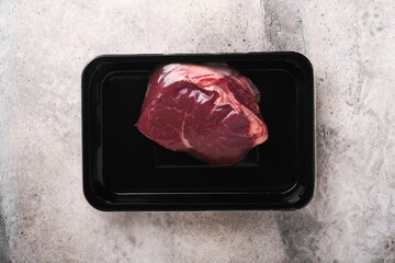 Beef steak. Fresh raw beef steak with fork and rosemary in vacuum black plastic pack on grey stone background. Vacoom package. Top view. Mockup for design idea.