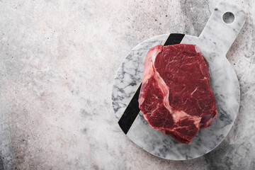 Raw beef steak. Fresh beef rib eye steak with fork rosemary, salt and pepper on marble stand on grey stone background. Top view. Mockup for design idea.