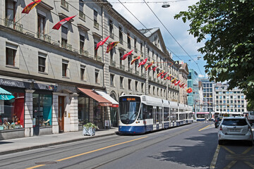 A tram passes through the rue de la Corraterie whose buildings are decked with the flags of Geneva...