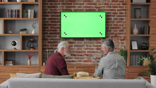 Fathers day. Adult son with senior father watching chroma-key green screen on tv, looking TV show or news, talking to each other and drinking beer. Two generations family enjoy spending time together.