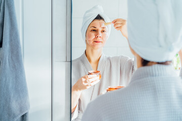 Woman in bathrobe with a towel on head looking in the mirror and applying facial cosmetic clay mask...