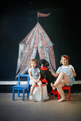 Obraz na płótnie Canvas Children together with pet dog play in theater, circus. Black poodle and European children against background of black chalk board with circus pattern.
