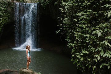 Woman stand and look at pool under falling water of Suwat waterfall in tropical jungle. Nature day tour, hiking activity adventure and fun at family tourist camp on summer vacation in Bali island