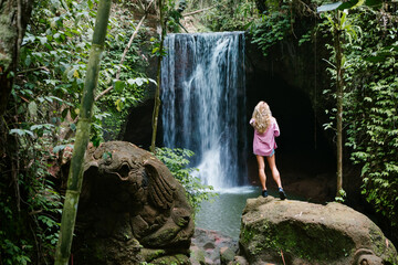 Fototapeta na wymiar Woman stand and look at pool under falling water of Suwat waterfall in tropical jungle. Nature day tour, hiking activity adventure and fun at family tourist camp on summer vacation in Bali island