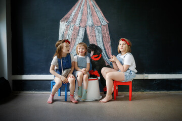 Fototapeta na wymiar Children together with pet dog play in theater, circus. Black poodle and three European kids against background of black chalk board with circus pattern.