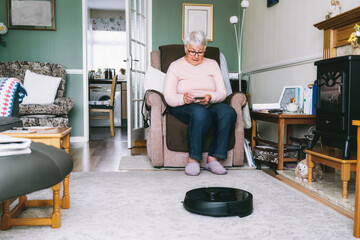 Senior woman using a robot vacuum cleaner through phone settings while sitting on armchair at home. Mature older lady holding mobile phone and control cleaning. Smart home. Seniors and technology. - obrazy, fototapety, plakaty