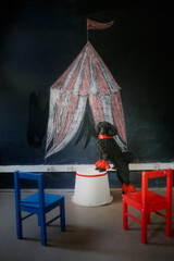 Black poodle dog in red outfit and collar sits at painted tent in front of chairs, dog before performance, playing circus