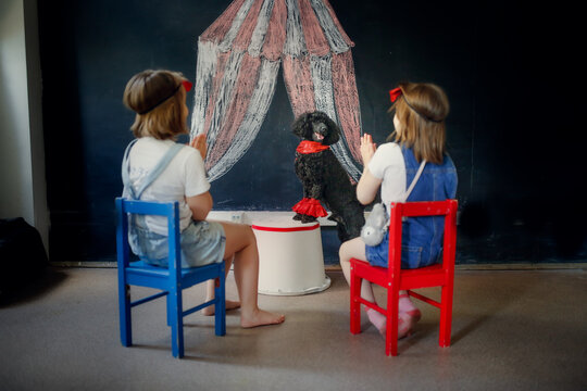 Two children watch poodle dog perform in front of painted circus, children clap their hands and rejoice in dog's performance. Black poodle is circus performer. Children and pet play circus.
