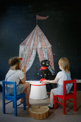 Two children watch poodle dog perform in front of painted circus, children clap their hands and rejoice in dog's performance. Black poodle is circus performer. Children and pet play circus.