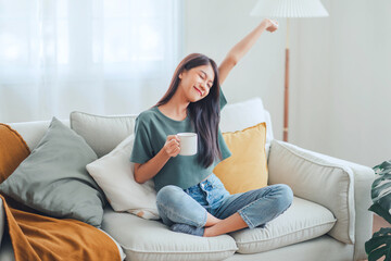 Happy young asian woman drinking coffee relaxing on sofa at home. Smiling female enjoying resting...