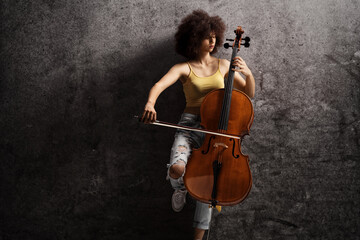 Young female artist playing a cello and leaning on a rusty wall - obrazy, fototapety, plakaty