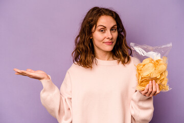 Young caucasian woman holding a bag of chips isolated on purple background showing a copy space on a palm and holding another hand on waist.