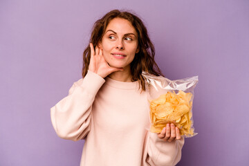 Young caucasian woman holding a bag of chips isolated on purple background trying to listening a gossip.