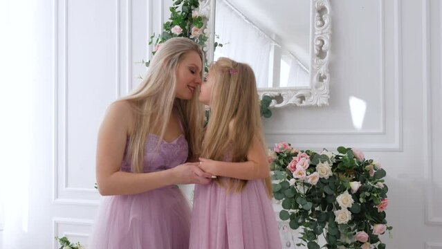 Happy young mom in purple dress with long blond hair hugging, kissing daughter in white room. Happy mother's day, 4k