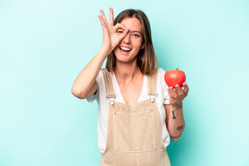 Young caucasian pregnant woman holding an apple isolated on blue background excited keeping ok...