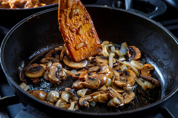 Mushrooms fried with onions in a deep frying pan with a wooden spatula.