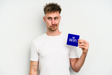 Young caucasian man holding wifi placard isolated on white background confused, feels doubtful and unsure.