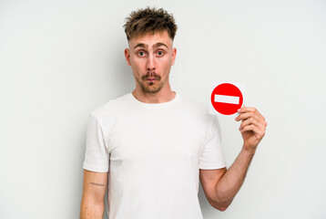 Young caucasian man holding a forbidden sign isolated on white background shrugs shoulders and open eyes confused.