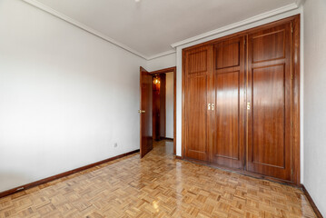 Empty room with built-in wardrobe with varnished wooden doors, oak parquet flooring and door to match the wardrobe