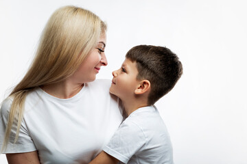 Fototapeta premium Mom and little son hug and smile. A beautiful blonde and a boy of 7 years old in white T-shirts look at each other. Love and tenderness. Light background.