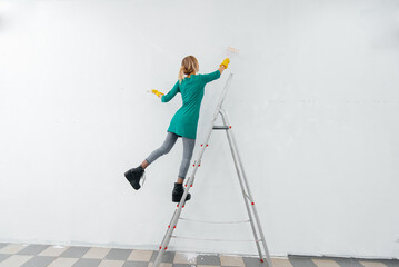 Obraz premium A young girl is engaged in repairs on the stairs and paints a white wall with a roller in a new apartment. Renovation of the interior and a new apartment. Housewarming and a desirable mortgage.