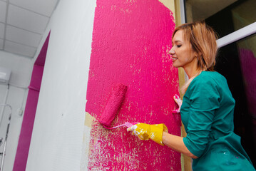 A young girl paints a wall with a roller in pink in her new apartment. Renovation of the interior and a new apartment. Housewarming and a desirable mortgage.