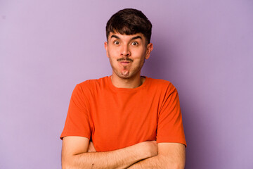 Young caucasian man isolated on purple background blows cheeks, has tired expression. Facial expression concept.