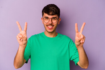 Young caucasian man isolated on purple background showing victory sign and smiling broadly.