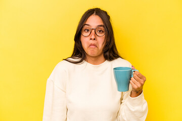 Young hispanic woman holding a mug isolated on yellow background shrugs shoulders and open eyes confused.