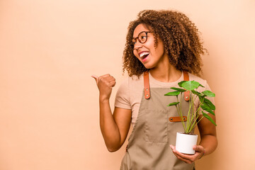 Young gardener African American woman holding plant isolated on beige background points with thumb...