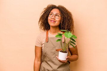 Young gardener African American woman holding plant isolated on beige background dreaming of...