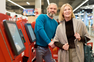 Happy aged couple in smart casualwear standing by self service terminals and looking at camera against cart with chosen food products