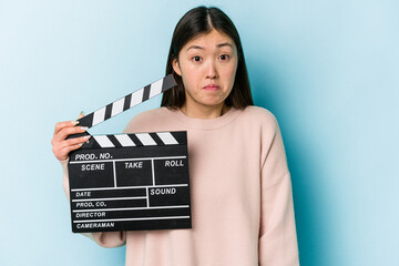 Young asian woman holding clapperboard isolated on blue background shrugs shoulders and open eyes...