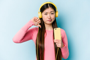Young asian woman listening to music isolated on blue background showing a dislike gesture, thumbs down. Disagreement concept.