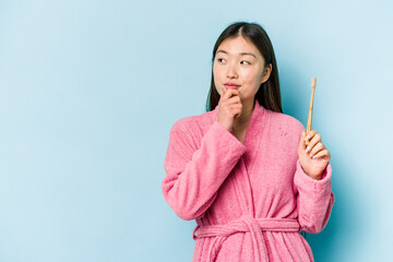 Young asian woman wearing a bathrobe brushing teeth isolated on pink background looking sideways...