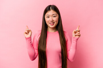 Young Chinese woman isolated on pink background indicates with both fore fingers up showing a blank...