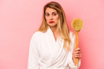Young caucasian woman holding back scratcher isolated on pink background shrugs shoulders and open...