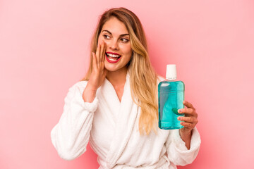 Young caucasian woman holding mouthwash wearing bathrobe isolated on pink background shouting and holding palm near opened mouth. - obrazy, fototapety, plakaty