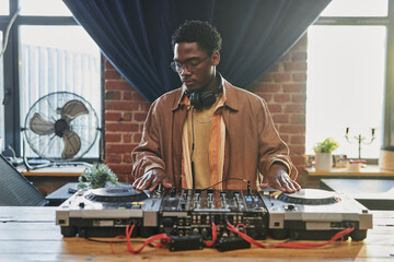Young black man in casualwear looking at dj board while touching turntables and adjusting musical...