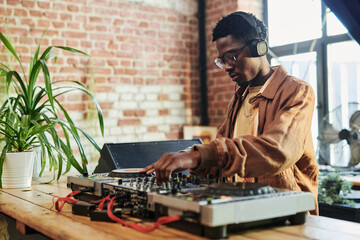 Serious young black man with headphones on his head mixing sounds while standing by turntables and making music in studio - obrazy, fototapety, plakaty