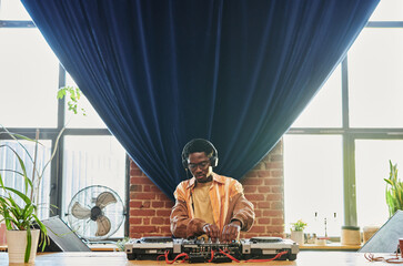 Young man in headphones and casualwear standing by table with dj set and turning mixers on console...