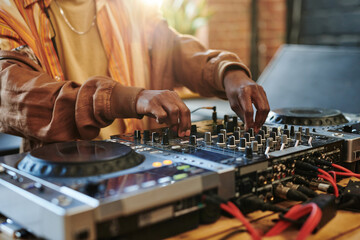 Hands of modern black man regulating sounds on dj set while standing by table in studio or loft apartment and turning mixers - obrazy, fototapety, plakaty