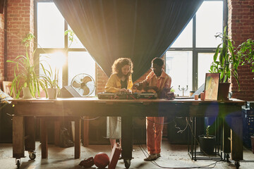 Young couple in casualwear standing by large wooden table with dj booth and creating new music...